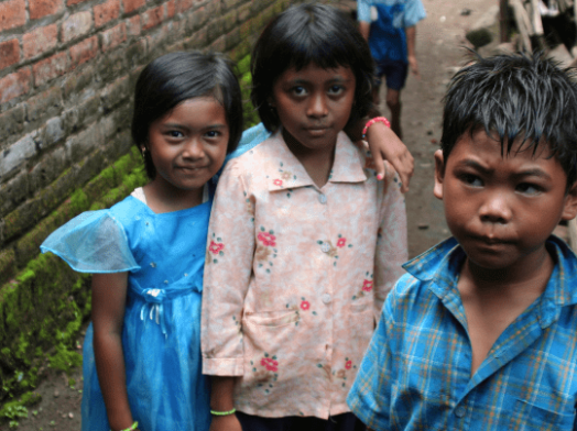 Three children standing in a local village in Indonesia, with traditional village surroundings.